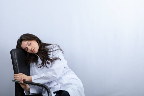 Portret lekarki w masce medycznej i białym płaszczu śpiącej na krześle. Wysokiej jakości zdjęcie - Zdjęcie, obraz