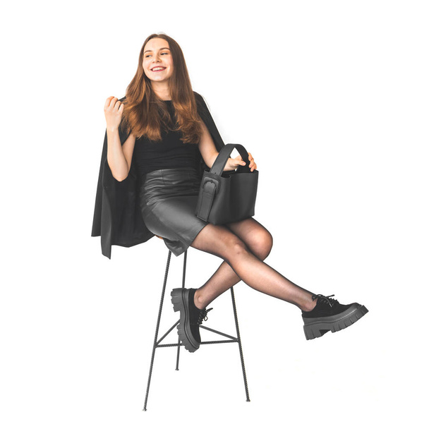 Glückliches junges Mädchen sitzt auf einem Stuhl, trägt schwarzes Kleid mit schwarzer Ledertasche, sexy Beine mit Stiefeln, Geschäftsfrau Konzept isoliert auf weißem Hintergrund - Foto, Bild