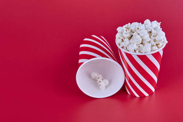 Two red glasses of popcorn on a red background. One stands full of popcorn, the other lies empty. - Foto, Bild