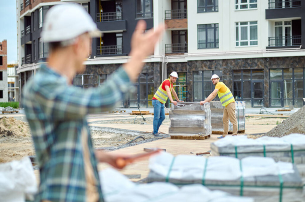 Two young builders looking at their colleague waving at them - 写真・画像