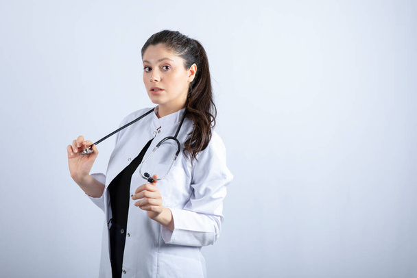 Beautiful doctor in white coat standing and posing over white wall. High quality photo - 写真・画像