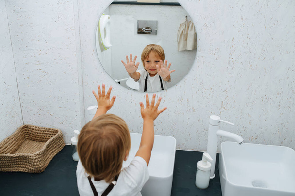 Smiling little boy washing his hands in the bathroom sink, waving clean palms at a mirror. From behind his shoulder. - Φωτογραφία, εικόνα