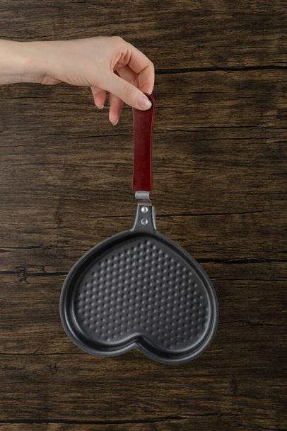 Woman hand holding an empty heart shaped frying pan on a wooden background. High quality photo - Photo, Image