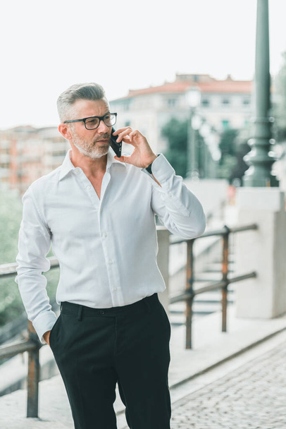 Businessman in good mood is talking on cellphone on his way to office. Handsome cheerful manager in formal suit walking enjoying getting to job in morning by foot having mobile conversation. Business communication concept - Photo, Image