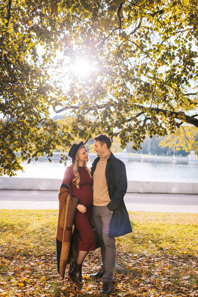 A couple of beautiful lovers spend time outdoors on the fall day. Pretty woman in a red dress and modish hat and a handsome man in a raincoat posing at the city park on a sunny autumn day - 写真・画像