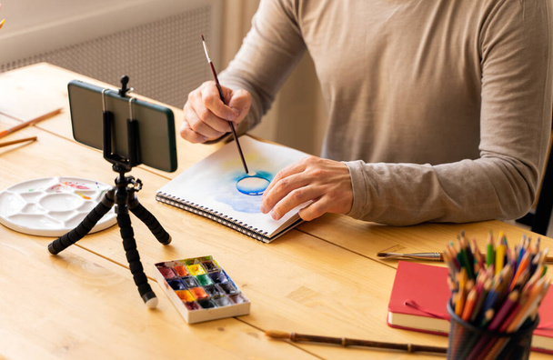 Stüdyoda öğrenciler için online resim dersi veren sanatçı erkek. Elinde boya fırçası, suluboya resim tutuyordu. Uzaktan kumanda öğrenimi kavramı. Uzayı kopyala Yatay. - Fotoğraf, Görsel