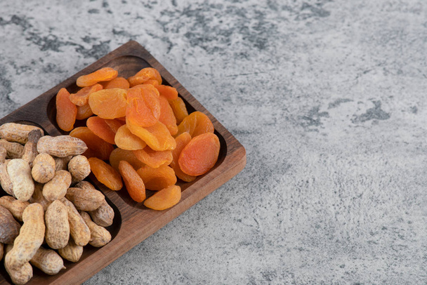 Wooden plate full of peanuts and dried apricots on marble surface. High quality photo - Photo, Image