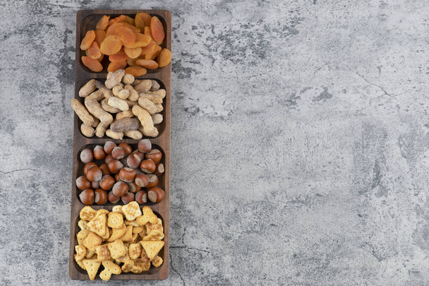 Holzteller mit verschiedenen Nüssen, Crackern und getrockneten Aprikosen auf Marmoroberfläche. Hochwertiges Foto - Foto, Bild