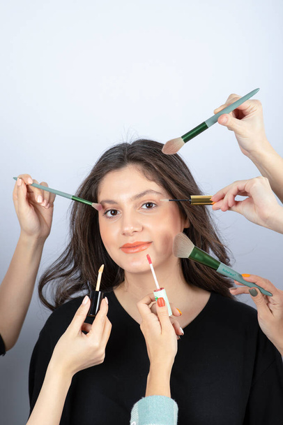 Smiling girl surrounded by hands of makeup artists with brushes, lipstick and mascara near her face. High quality photo - Photo, Image