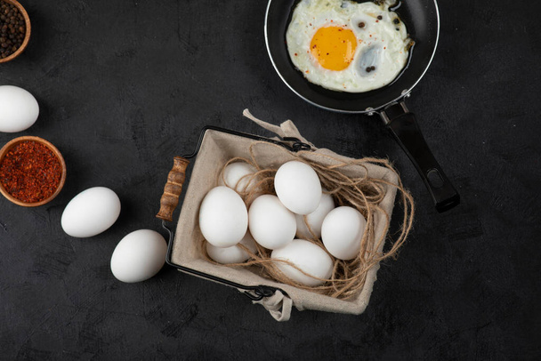 Pan smażone jajka, przyprawy i kosz surowych jaj na czarnym tle. Wysokiej jakości zdjęcie - Zdjęcie, obraz