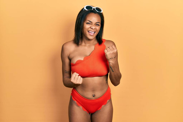 Junge afrikanisch-amerikanische Mädchen im Bikini feiern überrascht und erstaunt über den Erfolg mit erhobenen Armen und geschlossenen Augen. Siegerkonzept.  - Foto, Bild