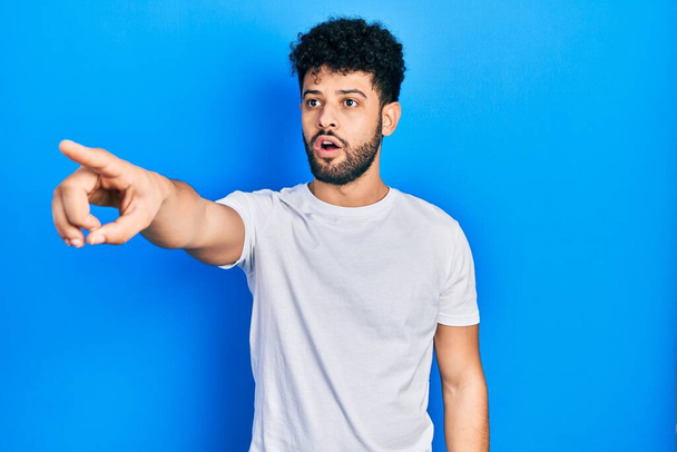 Young arab man with beard wearing casual white t shirt pointing with finger surprised ahead, open mouth amazed expression, something on the front  - Fotografie, Obrázek
