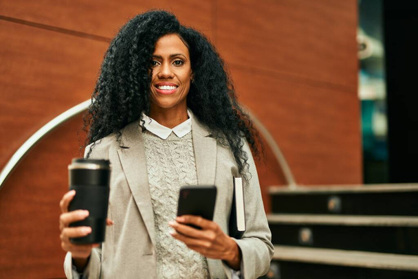 Middle age african american businesswoman using smartphone and drinking coffee at the city. - Photo, Image