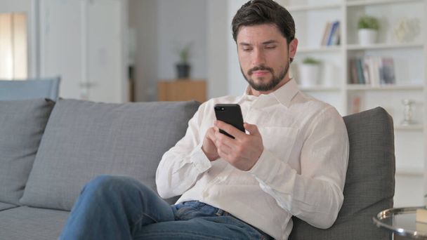 Houkutteleva nuori mies käyttää älypuhelinta kotona  - Valokuva, kuva