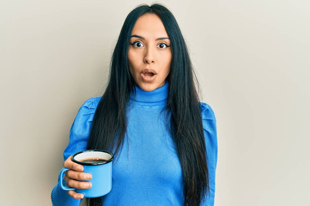 Молода іспаномовна дівчина тримає каву наляканою і здивована відкритим ротом для несподіванки, обличчя віри
  - Фото, зображення
