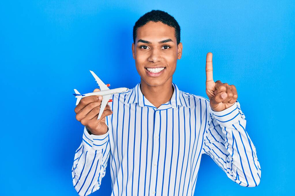 Joven afroamericano chico sosteniendo avión juguete sonriendo con una idea o pregunta señalando el dedo con la cara feliz, número uno  - Foto, Imagen
