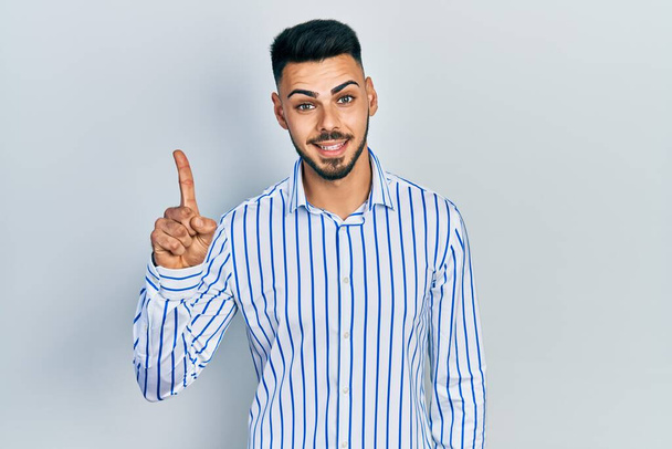Молодой латиноамериканец с бородой в обычной полосатой рубашке показывает и показывает пальцем номер один, улыбаясь уверенно и счастливо.  - Фото, изображение
