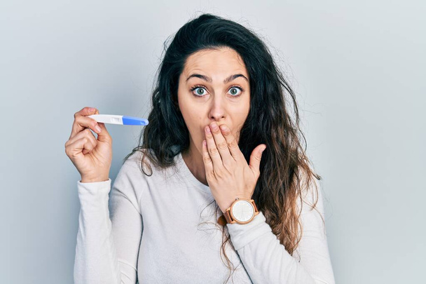Jonge Spaanse vrouw met een thermometer die haar mond met de hand bedekt, geschokt en bang voor fouten. verraste uitdrukking  - Foto, afbeelding