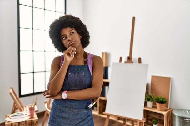 Młoda Afroamerykanka z afro-włosami w studiu sztuki z ręką na podbródku myśląca o pytaniu, zamyślonym wyrazie twarzy. Uśmiechając się z troskliwą twarzą. koncepcja wątpliwości.  - Zdjęcie, obraz
