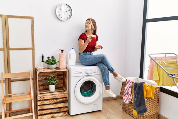 Young caucasian girl drinking coffee waiting for laundry sitting on whasing machine at home. - Photo, Image