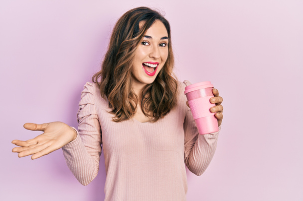 Joven chica hispana sosteniendo café celebrando logro con sonrisa feliz y expresión ganadora con la mano levantada  - Foto, imagen