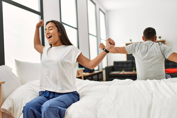 Young latin couple yawning and stretching arms sitting on the bed. - Photo, Image