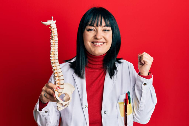 Jeune médecin hispanique femme tenant le modèle anatomique de colonne vertébrale criant fier, célébrant la victoire et le succès très excité avec le bras levé  - Photo, image