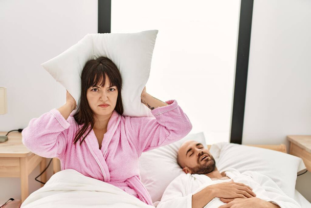 Spaanse vrouw boos met kussen op zijn oren terwijl de man snurkt op het bed. - Foto, afbeelding