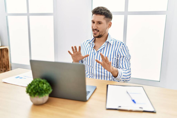 Young handsome man with beard working at the office using computer laptop afraid and terrified with fear expression stop gesture with hands, shouting in shock. panic concept.  - Photo, Image
