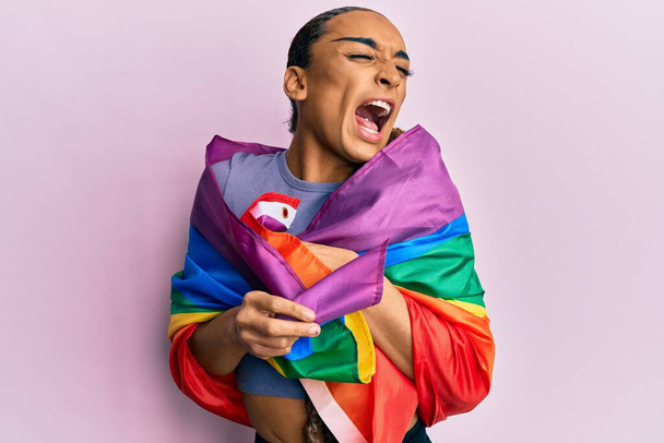 Hispanic man wearing make up and long hair wrapped in rainbow lgbtq flag angry and mad screaming frustrated and furious, shouting with anger. rage and aggressive concept.  - Photo, Image