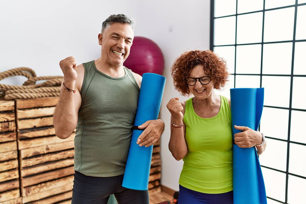 Middle age couple holding yoga mat very happy and excited doing winner gesture with arms raised, smiling and screaming for success. celebration concept.  - Foto, afbeelding