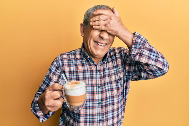 Красивый пожилой мужчина с седыми волосами пьет чашку кофе, улыбаясь и смеясь, закрывая глаза рукой. слепая концепция.  - Фото, изображение