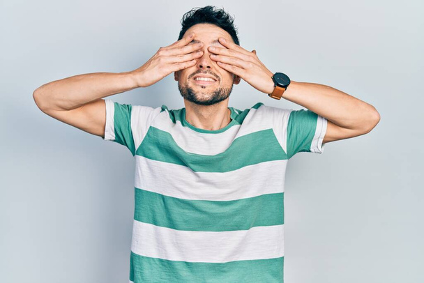 Young hispanic man wearing casual clothes covering eyes with hands smiling cheerful and funny. blind concept.  - Photo, Image