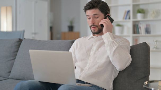 Młody człowiek z laptopem rozmawia przez telefon w domu  - Zdjęcie, obraz