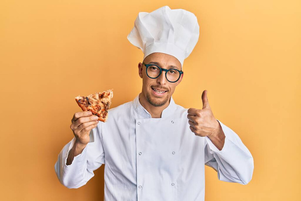 Лысый мужчина с бородой носит профессиональный фартук повара держа итальянскую пиццу улыбаясь счастливый и позитивный, большой палец вверх делает отлично и знак одобрения  - Фото, изображение