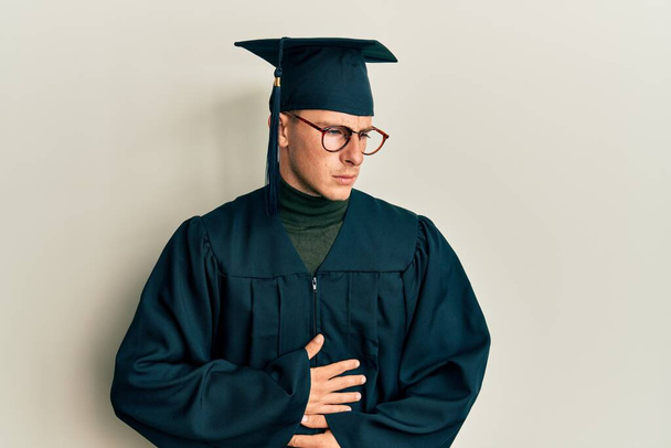 Young caucasian man wearing graduation cap and ceremony robe with hand on stomach because indigestion, painful illness feeling unwell. ache concept.  - Fotó, kép