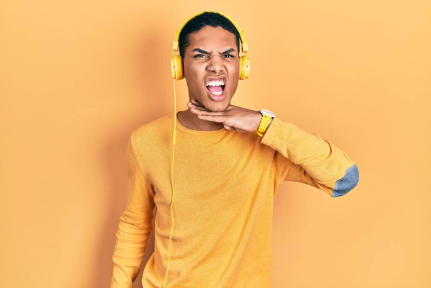 Νεαρός Αφροαμερικάνος που ακούει μουσική με ακουστικά να κόβουν το λαιμό με το χέρι ως μαχαίρι, απειλεί την επιθετικότητα με έξαλλη βία  - Φωτογραφία, εικόνα