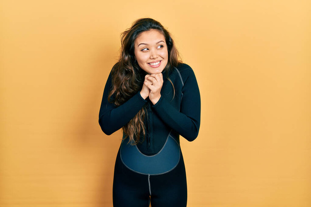 Молодая испаноязычная девушка в форме водолаза неопрена смеется нервно и взволнованно с руками на подбородке, глядя в сторону  - Фото, изображение