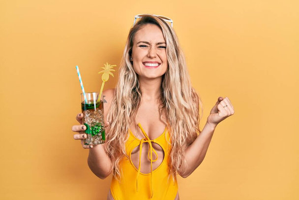 Mooie jonge blonde vrouw draagt badpak drinken tropische cocktail schreeuwen trots, vieren overwinning en succes zeer enthousiast met opgeheven arm  - Foto, afbeelding