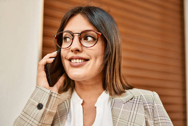 Young hispanic businesswoman smiling happy talking by the smartphone at the city. - 写真・画像