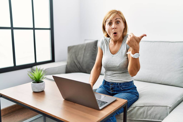 Blonde Frau mittleren Alters mit Laptop zu Hause überrascht mit Zeigefinger zur Seite, offener Mund erstaunter Ausdruck.  - Foto, Bild