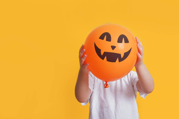 funny child in a white t-shirt holding an orange balloon with a picture of jack lantern, halloween mock up, yellow background copy space - Foto, Imagem