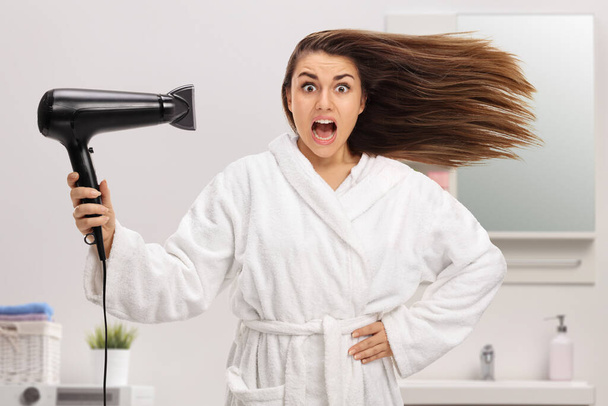 Mujer joven sorprendida en un albornoz secador de pelo en un baño - Foto, imagen