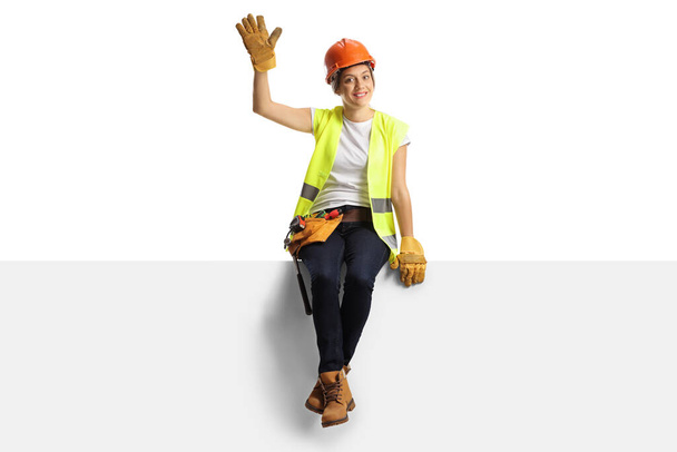 Full length portrait of a cheerful female engineer sitting on a blank panel and waving isolated on white background - Foto, imagen