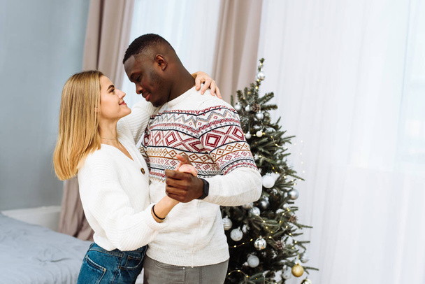 Joven hermosa pareja de amantes están bailando, mirándose a los ojos junto al árbol de Navidad. Hermosa joven pareja enamorada - Foto, Imagen