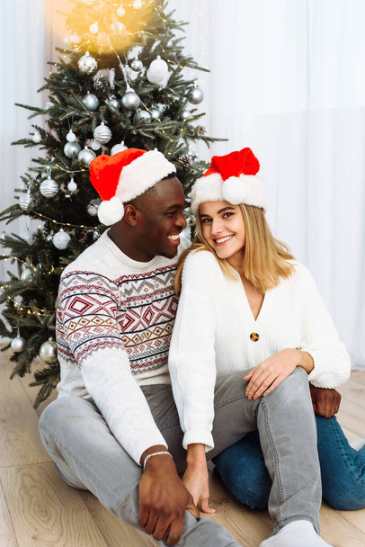 Portrait of a couple in love in santa hats sitting by the tree. Glare of the yellow garland. Black male hugs a Caucasian woman - Zdjęcie, obraz