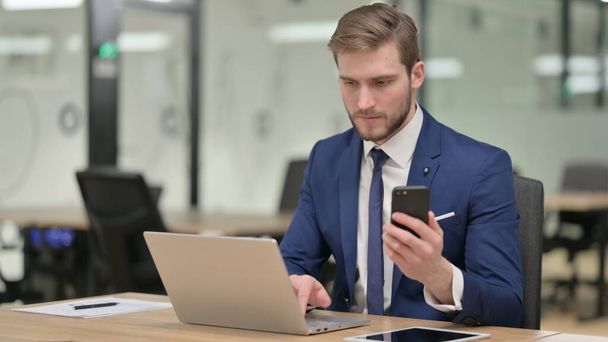 Biznesmen z laptopem przy użyciu smartfona w pracy - Zdjęcie, obraz