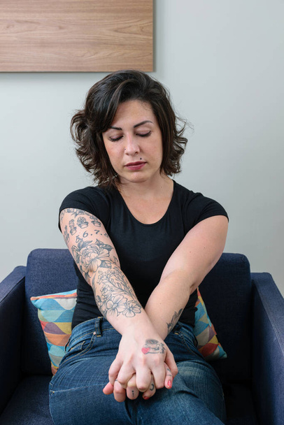Mujer brasileña, tatuada, con las manos cruzadas y mirando hacia abajo, mostrando preocupación y ansiedad. - Foto, Imagen