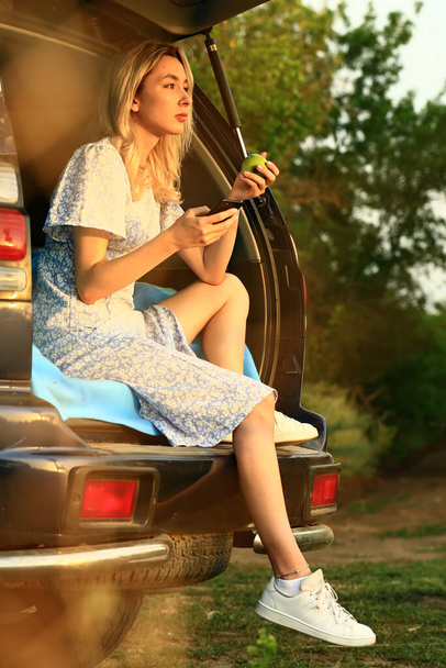 νεαρή γυναίκα κάθεται στο ανοιχτό πορτ-μπαγκάζ ενός αυτοκινήτου και τρώει ένα πράσινο μήλο. - Φωτογραφία, εικόνα