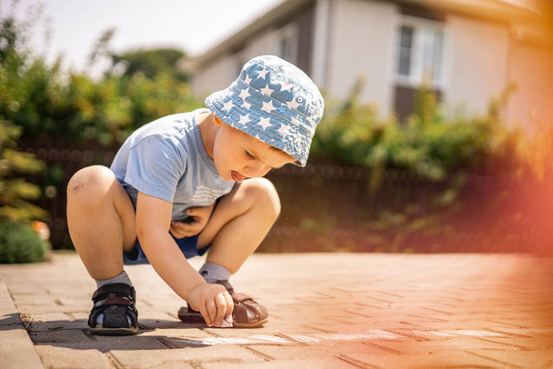 Cheerful little boy drawing hopscotch with chalk on ground enjoying happy childhood at backyard - Photo, Image